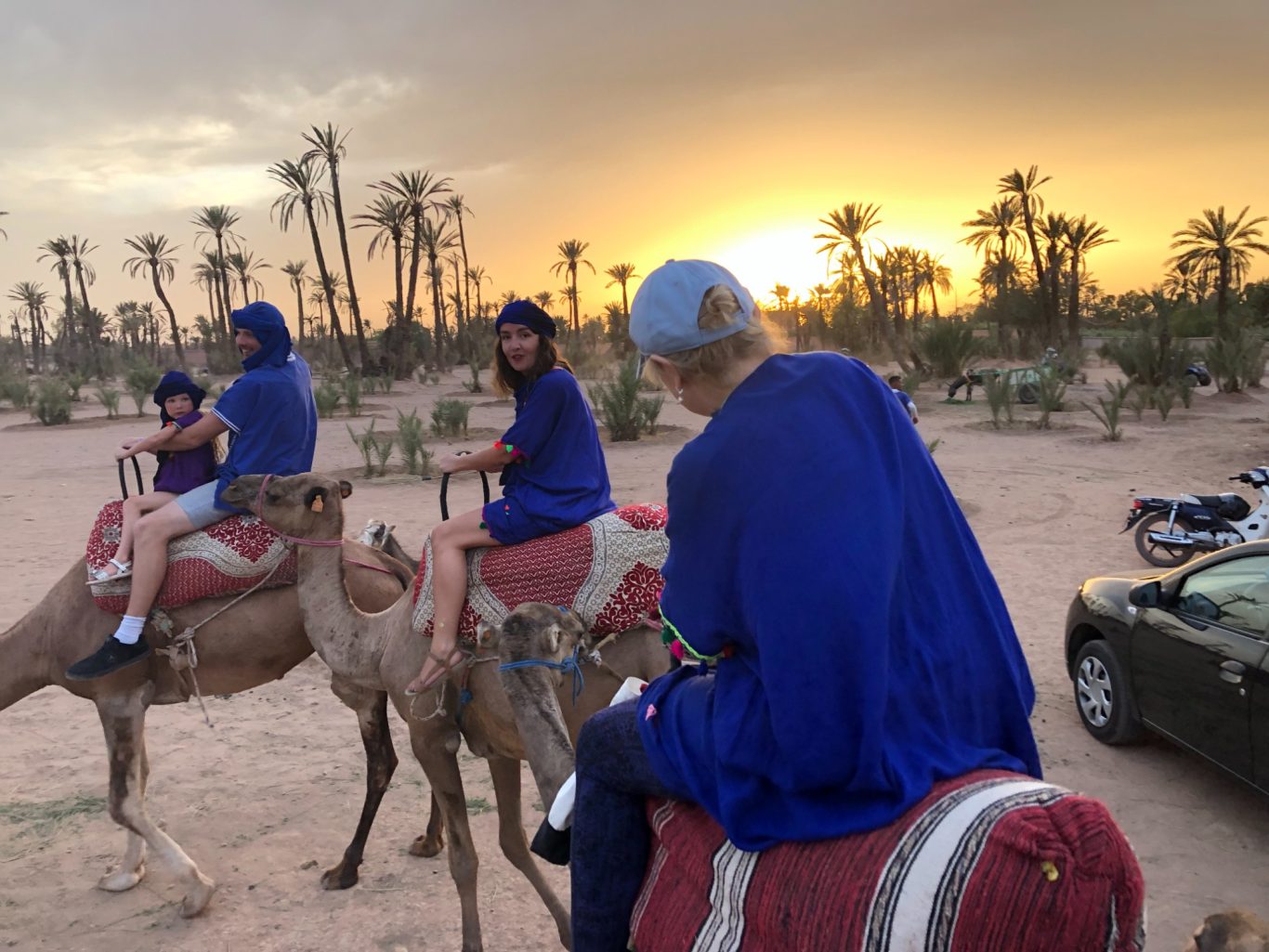 People riding camels in a desert landscape at sunset with palm trees in the background.