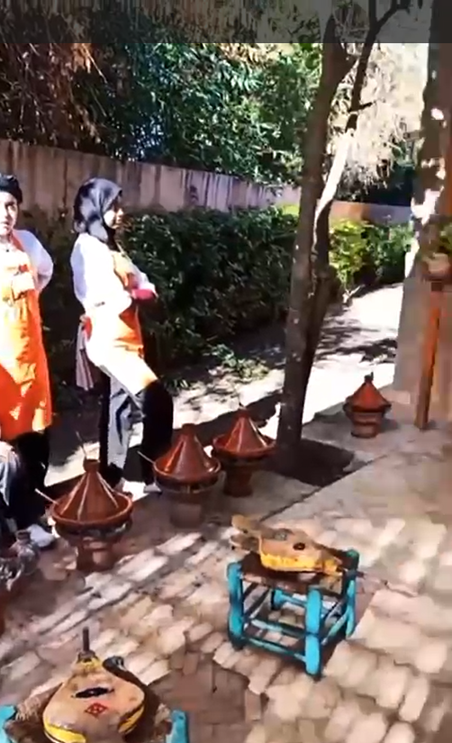Two people in aprons stand next to traditional cooking pots outdoors.