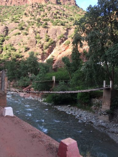 A wooden suspension bridge crosses a river, surrounded by lush greenery and rocky hills.