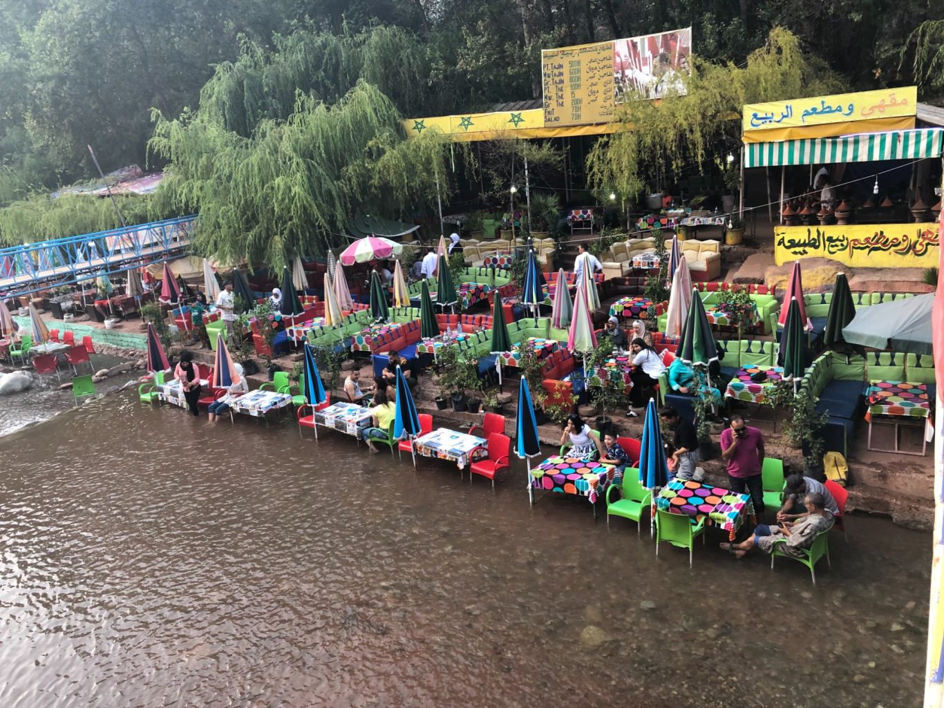 Ourika valley A bustling riverside market with colourful stalls and people seated by the water.